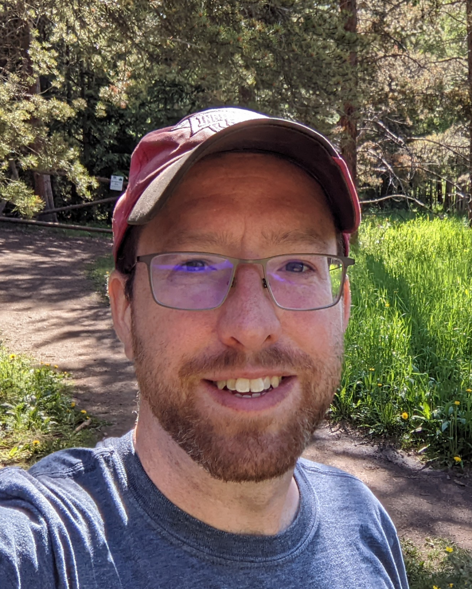 Smiling man in a baseball hat and t-shirt on a path in the grass with woods in the background