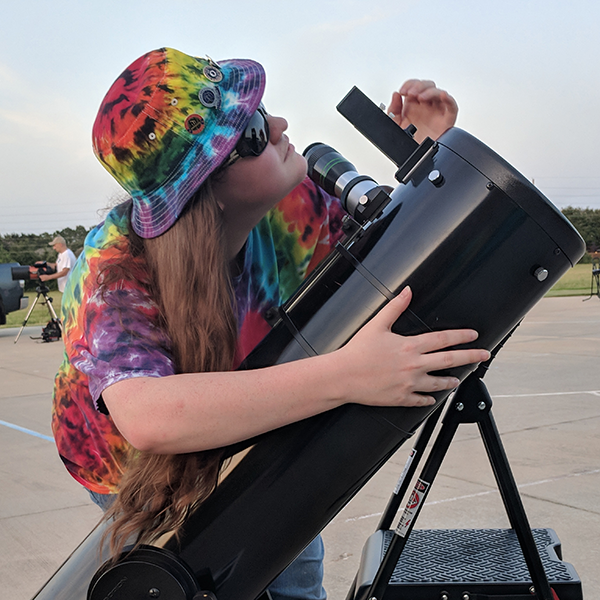 A young woman in a tie-dyed hat and shirt looking through a telescope with her arm on it, standing on pavement.